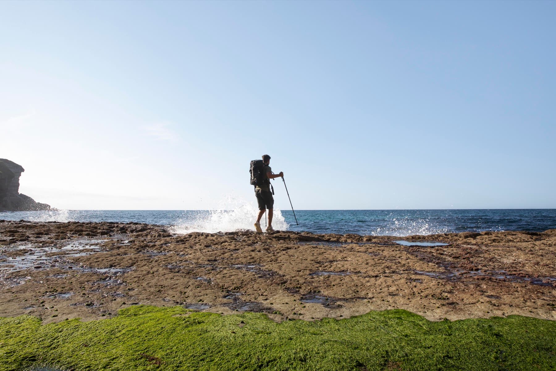 Person standing on a rocky shoreline platform with a pole, waves breaking behind them